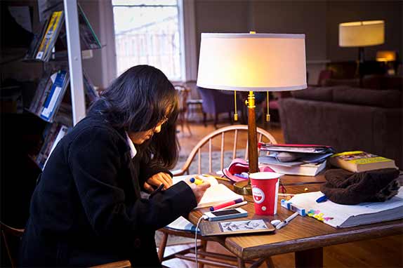 student studying by lamplight with coffee cup, books, and bookbag on table